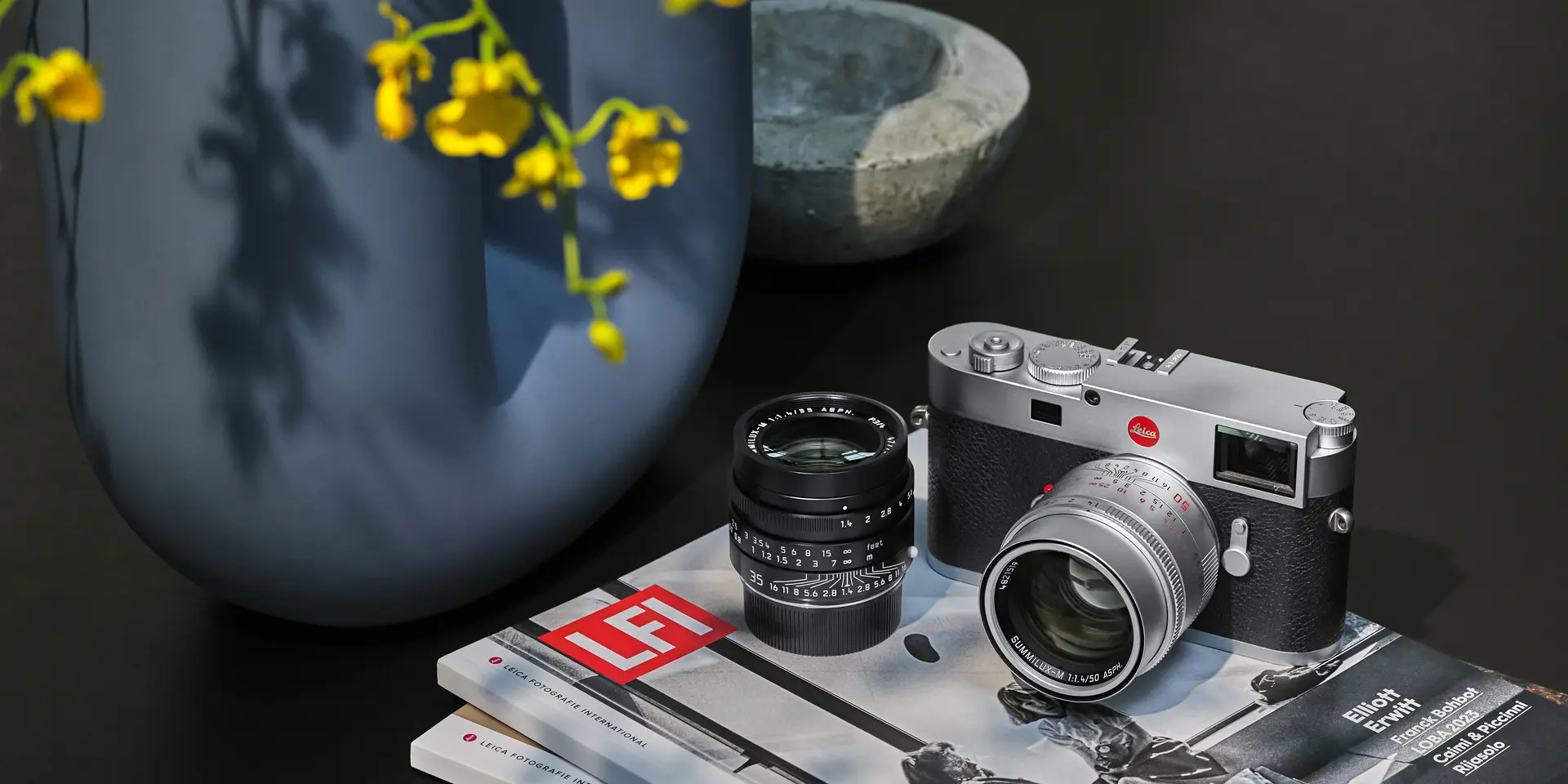Leica camera and lens on top of two books, with a vase of flowers in the background.